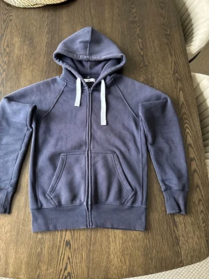 Blå zip hoodie med vita snören - Mysig blå hoodie med dragkedja och vita snören i huvan. Tröjan har känguruficka framtill och ribbade muddar vid ärmar och nederkant. Perfekt för chill dagar eller när du vill ha en enkel och stilren look.