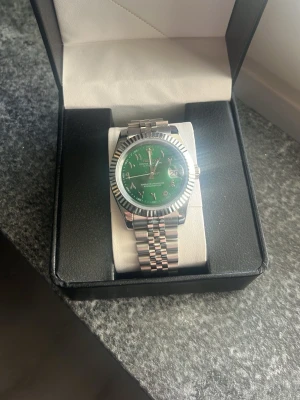Seiko custom mod Datejust (Arabic) - Säljer en oanvänd Seiko Custom Mod Datejust 39 mm med en slående grön urtavla, arabiska siffror och ett klassiskt Jubilee-armband. Klockan har ett automatiskt urverk och är tillverkad i stål. Den är i oanvänt skick då jag själv inte kan så mycket om klockor och därför inte vill säga mer än så. Fick den som present men har aldrig använt den! Pris kan diskuteras.