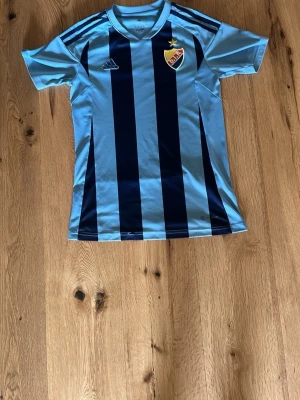 Ljusblå Djurgården matchtröja - Säljer en ljusblå och mörkblå randig matchtröja från Djurgården med klubbmärke på bröstet och Adidas-logga. Tröjan har korta ärmar och klassiska Adidas-ränder på axlarna. Perfekt för fotbollsträning eller att bära på matchdag.