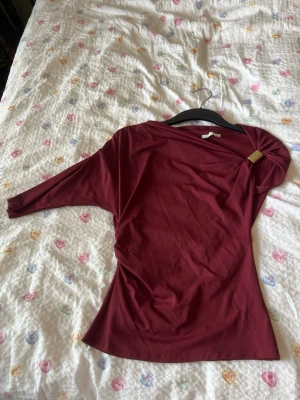 Vinröd oneshoulder topp - Säljer en vinröd topp med oneshoulder-design och lång ärm. Toppen har en mjuk och stretchig känsla, perfekt för dig som gillar en stilren och modern look. Passformen är slim och materialet känns som polyester.