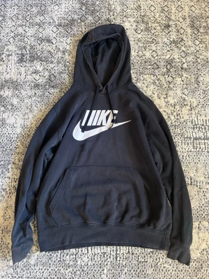 Svart hoodie från Nike med logga - Säljer en svart hoodie från Nike med stor vit logga och swoosh på bröstet. Hoodien har huva med dragsko och en stor magficka. Perfekt för dig som gillar streetwear och sportig stil. Materialet känns mjukt och bekvämt.