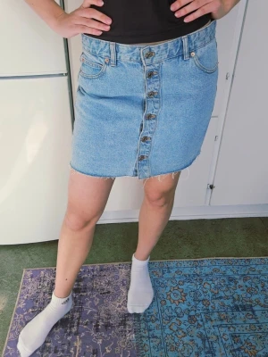 Ljusblå jeanskjol med knappar st XL  - Säljer en ljusblå jeanskjol i kort modell med rå kant nertill och knappar framtill. Kjolen har klassiska fickor fram och bak samt bälteshällor. Perfekt för dig som gillar en enkel men snygg denimlook. Ej retur.