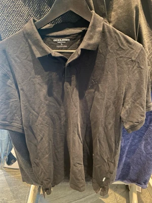 Svart pikétröja från Jack & Jones - Klassisk svart pikétröja från Jack & Jones med krage och knappar framtill. Tillverkad i mjuk bomull och har korta ärmar. Perfekt till jeans eller shorts för en clean och enkel stil.