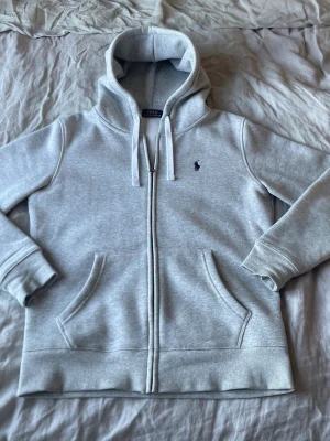 Ralph Lauren Hoodie - Ljusgrå hoodie med dragkedja från Polo Ralph Lauren. Klassisk design med huva, vita dragsnören och broderad logga på bröstet. Mjuk bomullsblandning och två fickor framtill. Perfekt för en avslappnad stil.