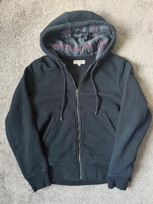Burberry zip hoodie  - Snygg mörkblå hoodie med dragkedja och huva. Insidan av huvan är fodrad med grått tyg och har rosa detaljer. Praktiska fickor framtill och ribbade muddar vid ärmar och nederkant. Perfekt för chill dagar eller när du vill ha något bekvämt. Vid frågor eller funderingar hör av dig 😁🙌