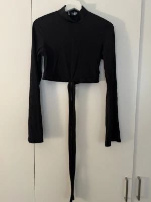 Svart croppad långärmad topp - Snygg svart croppad topp med lång ärm och hög krage. Toppen har långa knytband framtill som ger en cool detalj. Perfekt att styla med höga jeans eller kjol för en trendig look.
