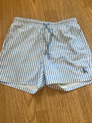 Randiga badbyxor från Calvin Klein - Snygga badbyxor från Calvin Klein med blå och vita vertikala ränder. De har resår i midjan med dragsko, sidofickor och en bakficka. Lätta och snabbtorkande, perfekta för stranden eller poolen. CK-logga broderad på benet.