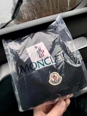 Moncler mössa  - Snygg svart ribbstickad mössa från Moncler med klassisk Moncler-logga framtill. Mössan har uppvikt kant och ar tillverkad i ett mjukt och varmande material, perfekt för kyliga dagar. En stilren accessoar som lyfter din outfit. Tag och äkthetsmärke finns kvar.