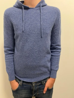 Blå Kashmir hoodie  - Säljer en blå hoodie med huva och dragsnören. Tröjan är gjord i en mjuk och lite luddig ullmix som känns riktigt skön mot huden. Långärmad modell med ribbade muddar vid ärmslut och nederkant. Perfekt för dig som gillar en enkel men stilren look.lite knopprig fixar såkalrt de innan ja skickar den