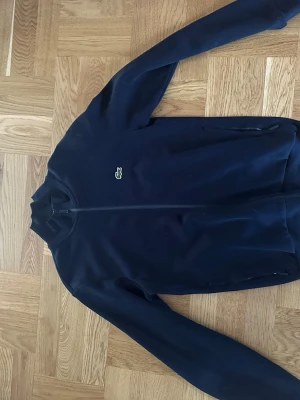 Mörkblå ziptröja från Lacoste - Snygg mörkblå ziptröja från Lacoste med klassisk krokodillogo på bröstet. Tröjan har hel dragkedja framtill, ståkrage och två fickor med dragkedja. Perfekt för en avslappnad och sportig stil.