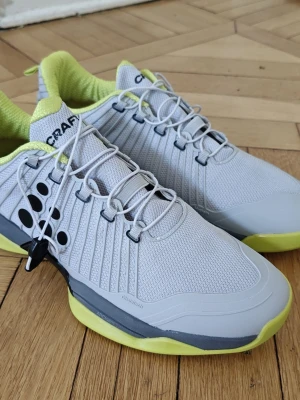Craft i1 cage sneakers i grått och neongult 41,5 - Snygga sneakers från Craft i ljusgrå mesh med neongula detaljer och svart logotyp. Skorna har lågt skaft, snörning och en sportig design med mönstrad sula i gult och orange. Perfekta för dig som gillar en modern och färgstark look. Skorna är helt nya och aldrig använda. Deras Etiketterna är borta för att skilja dem från dem använda tvillingarna . 