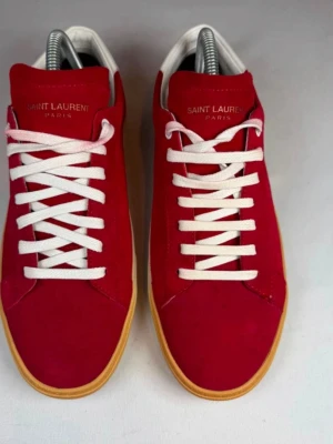 Röda Saint Laurent sneakers i mocka - Snygga röda sneakers från Saint Laurent i mjuk mocka med vita skosnören och beige gummisula. Klassisk låg modell med stilren design och diskret logga på plösen. Perfekta för dig som vill ha en clean och trendig look. Använd 1 gång bara
