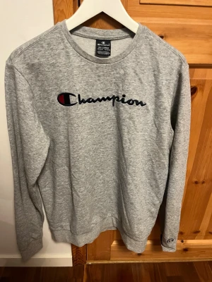 Grå sweatshirt från Champion - Snygg grå sweatshirt från Champion med klassisk logga broderad i svart, vitt och rött på bröstet. Tröjan har rund halsringning, långa ärmar och mjukt material som passar perfekt till en avslappnad stil.
