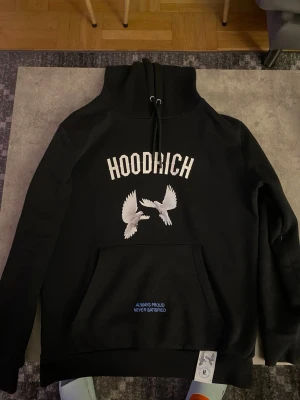 Svart hoodie från Hoodrich med duvor - Svart hoodie från Hoodrich med stor vit logga och två broderade duvor på bröstet. Klassisk känguruficka framtill och texten 'ALWAYS PROUD NEVER SATISFIED' på fickan. Justerbar huva med snören och ribbade muddar.