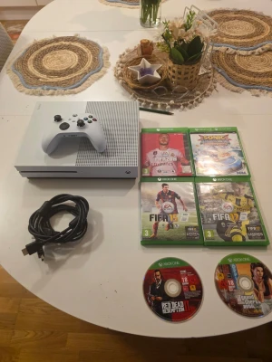 Xbox One S med spel och handkontroll - Säljer en vit Xbox One S med en matchande vit handkontroll. Medföljer strömkabel och flera spel, bland annat FIFA 17, FIFA 15, FIFA 20, Sonic Mania, Red Dead Redemption II och GTA V. Konsolen har en stilren design i plast och passar perfekt för gaming.