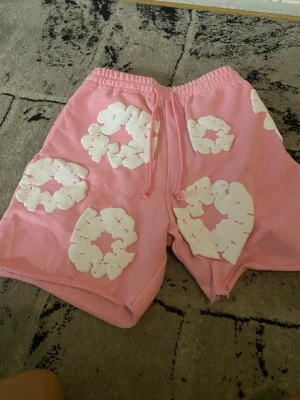 Rosa shorts med vita blommor - Säljer ett par rosa shorts med vita blommönster och snörning i midjan. Shortsen har en avslappnad passform och är perfekta för varma dagar. De är tillverkade i mjukt material och har elastisk midja för extra komfort.