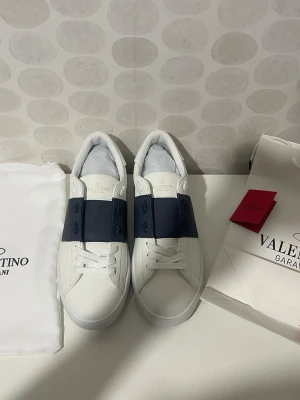 Vita sneakers med blå detalj Valentino - Snygga vita sneakers från Valentino med bred mörkblå rem över snörningen. Skorna har klassisk låg profil, rund tå och är tillverkade i slätt läder. Perfekta för dig som vill ha en clean och stilren look med exklusiv känsla.