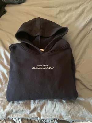 Svart hoodie med Stockholm-tryck - Säljer en svart hoodie med texten 'Notre succès' och stort 'STOCKHOLM Private Club' tryckt på bröstet. Hoodien har huva och är gjord i mjukt material, perfekt för en chill och stilren look. Passar dig som gillar streetwear och enkel design.