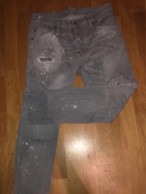 Dsquared jeans - Säljer ett par grå jeans med vita färgstänk och slitningar på ena låret. Jeansen har klassisk femficksmodell och raka ben. Perfekta för dig som gillar en lite mer edgy och avslappnad stil.