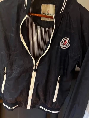 Svart vindjacka från Moncler - Snygg svart vindjacka från Moncler med vit dragkedja och vita detaljer vid fickorna. Jackan har Moncler-logga på bröstet och randigt innerfoder. Perfekt för dig som vill ha en stilren och sportig look.