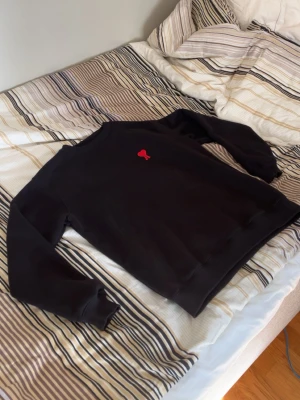 Ami Paris Tjocktröja - En svart sweatshirt med rund hals och en liten röd broderad detalj på bröstet. Tröjan har långa ärmar och ribbade muddar vid ärmslut och nederkant. Perfekt för en avslappnad och stilren look. Ami paris knappast använd.