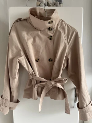 Trenchcoat från bershka  - Snygg beige trenchcoat i kort modell med dubbelknäppning, hög krage och knytskärp i midjan. Jackan har axelklaffar, dekorativa knappar och justerbara ärmslut. Perfekt för dig som vill ha en klassisk men trendig look.