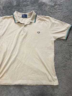Beige pikétröja från Fred Perry - Säljer en beige pikétröja från Fred Perry med klassisk krage och knappar. Tröjan har svarta och turkosa ränder på kragen och ärmsluten samt den ikoniska Fred Perry-loggan broderad på bröstet. Tillverkad i bomull och har en slim fit passform.
