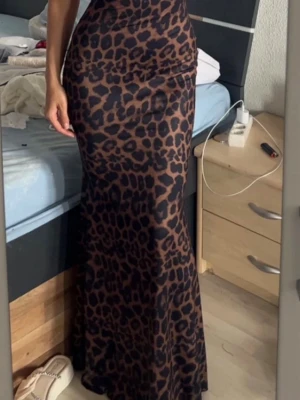 Lång leopardmönstrad klänning - Säljer en lång, figurnära klänning med leopardmönster i brunt och svart. Klänningen har tunna axelband och öppen rygg med snygga korsade banddetaljer. Materialet ser ut att vara stretchigt och följsamt, perfekt för dig som vill sticka ut.