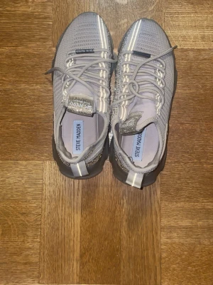 Grå Steve Madden sneakers med mesh - Snygga grå sneakers från Steve Madden med stickad mesh-överdel och reflekterande detaljer. Skorna har tjock sula och snörning framtill. Perfekta för en trendig och sportig look.