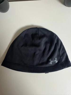 Svart mössa från Under Armour - Snygg svart mössa från Under Armour med diskret logga i silver på sidan. Tillverkad i ett stretchigt och mjukt material som sitter skönt på huvudet. Perfekt för kyliga dagar och sportiga outfits.