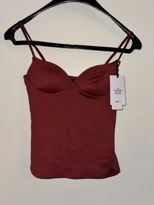Vinröd croppad linnetopp - Trendig vinröd linnetopp med smala axelband och vadderade kupor. Toppen är croppad och har en figurnära passform som framhäver siluetten. Perfekt att matcha med höga jeans eller kjol för en snygg look.
