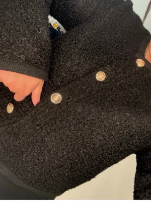 Kofta/jacka från gina med guldknappar - Stilren svart kofta i bouclématerial med dekorativa guldknappar framtill. Koftan har rund halsringning, långa ärmar och en mjuk, fluffig struktur som ger en lyxig känsla. Perfekt att slänga över en topp för en trendig look.