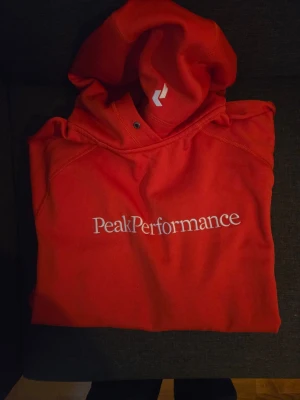  hoodie från Peak Performance - Snygg orange hoodie från Peak Performance med stor vit logga på bröstet. Klassisk modell med huva och dragsko, perfekt för en avslappnad och sportig stil. Tillverkad i mjukt material som känns skönt mot huden.