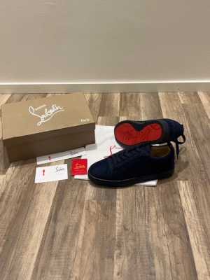 Mörkblå sneakers från Louboutin - Snygga mörkblå sneakers från Christian Louboutin med klassisk röd sula och diskret design. Skorna har ovandel i mocka, svarta skosnören och lågt skaft. Perfekta för dig som vill ha en stilren och exklusiv look.