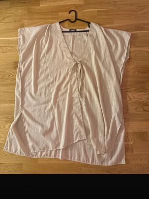 Beige beach cover Up med knyt - Luftig beige beach cover up med subtila ränder och knyt framtill. Plagget har v-ringning, ärmlös design och slits i sidorna. Perfekt för en avslappnad och trendig stil.
