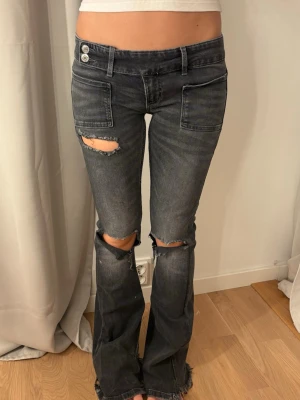 Grå slitna bootcut jeans med hål Young tall - Säljer ett par grå bootcut jeans med slitningar och hål vid knäna och låret. Jeansen har råa kanter nertill, två framfickor och två bakfickor samt dubbla knappar i midjan. Perfekta för en avslappnad och trendig look.