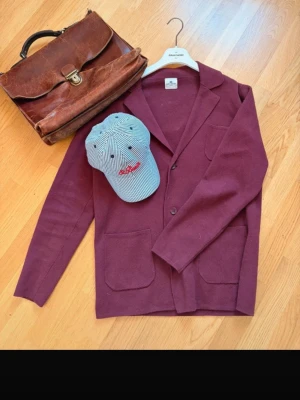 John Henric Burgundy Knit Blazer – 100% Cotton – Stl S - Snygg och stilren John Henric blazer/kofta i burgundy/vinröd färg, tillverkad i 100% bomull. Modellen har en elegant krage, tre knappar framtill och fyra praktiska utanpåliggande fickor.  Perfekt att bära över en skjorta för en smart casual / preppy / old money-look, men fungerar även mer avslappnat. Fin textur i materialet och en tidlös modell som passar både vår, sommar och höst.  Märke: John Henric Storlek: S Material: 100% Cotton Färg: Burgundy / vinröd / mörkröd