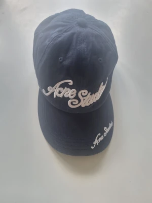 Mörkblå keps från Acne Studios - Snygg mörkblå keps från Acne Studios med vit broderad logga framtill och på skärmen. Klassisk böjd skärm och justerbar baksida. Tillverkad i 100% bomull för en bekväm och luftig känsla.