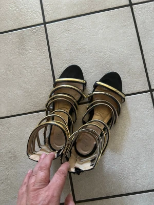 Svarta sandaler med gulddetaljer storlek 38 - Säljer ett par svarta sandaler med flera breda remmar dekorerade med guldfärgade detaljer. Sandalerna har öppen tå och klack, samt justerbar rem runt ankeln. Materialet ser ut att vara en mix av mockaimitation och syntet. Perfekta för dig som vill sticka ut.
