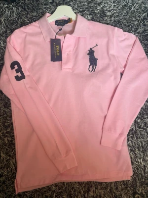 Rosa långärmad piké från Polo Ralph Lauren - Säljer en rosa långärmad pikétröja från Polo Ralph Lauren med klassisk krage och knappar. Stor mörkblå logga på bröstet och siffran 3 på ärmen. Perfekt för dig som gillar stilren och sportig look. Helt ny med lappar kvar storlek S