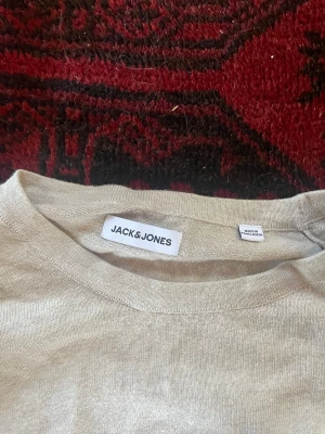 Beige tröja från Jack & Jones - En stilren beige tröja från Jack & Jones med rund halsringning och långa ärmar. Tröjan har ribbade muddar vid ärmslut och nederkant, vilket ger en snygg och enkel look. Perfekt att matcha med jeans eller chinos.