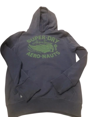 Blå Superdry hoodie med tryck - Mörkblå hoodie från Superdry med stort grönt tryck på bröstet där det står 'Superdry AeroSpace Aero-Nauts'. Klassisk känguruficka framtill och huva med dragsko. Tillverkad i mjukt bomullsmaterial, perfekt för en avslappnad stil.