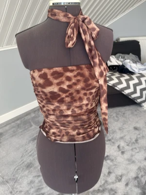 Brun leopardmönstrad halterneck tubtopp - Säljer en brun leopardmönstrad tubtopp med halterneck och knyt i nacken. Toppen är tight och croppad, perfekt för dig som vill sticka ut. Materialet känns mjukt och stretchigt, troligen polyester. Supersnygg till både jeans och kjol! (KÖPTE I THAILAND) aldrig använt 