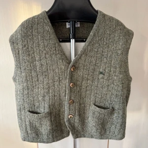 Burberry Grå stickad väst med knappar - Snygg gråmelerad stickad väst med ribbad struktur och v-ringning. Västen har fyra knappar framtill och två fickor. Perfekt att slänga över en skjorta för en chill look. 0374