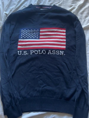Mörkblå tröja U.S. Polo Assn. - Mörkblå långärmad tröja från U.S. Polo Assn. med stor amerikansk flagga på bröstet och texten 'U.S. POLO ASSN.' under. Tröjan har ribbade muddar vid ärmslut och nederkant. Perfekt för dig som gillar klassisk och sportig stil.