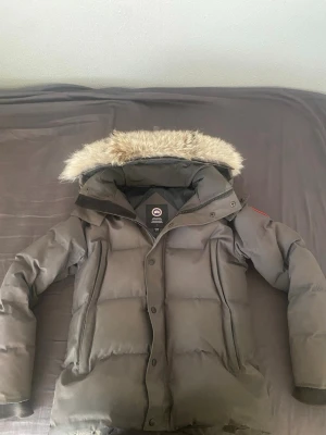 Grå dunjacka från Canada Goose - Säljer en svart dunjacka från Canada Goose med avtagbar huva och snygg pälskant. Jackan har en klassisk puffer-modell, stora fickor och diskret logga på ärmen. Perfekt för kalla vinterdagar och riktigt varm tack vare dunfyllningen.