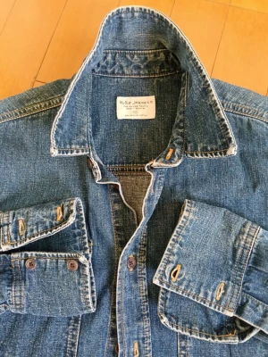 Jeansskjorta från Nudie Jeans - Klassisk blå jeansskjorta från Nudie Jeans med två bröstfickor och knappar framtill. Skjortan har lång ärm och är tillverkad i slitstarkt denimtyg av bomull. Perfekt för en avslappnad och cool stil.