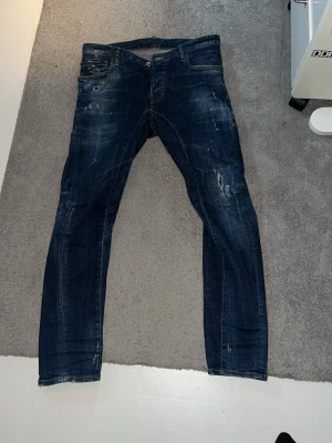 Mörkblå slim fit jeans med slitningar - Säljer ett par mörkblå slim fit jeans med coola slitningar och färgstänk. Jeansen har fem fickor, låg midja och detaljerade sömmar. Bakfickan har en röd DSC-logga. Perfekta för dig som gillar en streetwear-look. Passar 28-30 L32. Super fint skick på dom och är ärvt från en i min familj som ej passar i dom längre, köpta i butik för över 8 år sen
