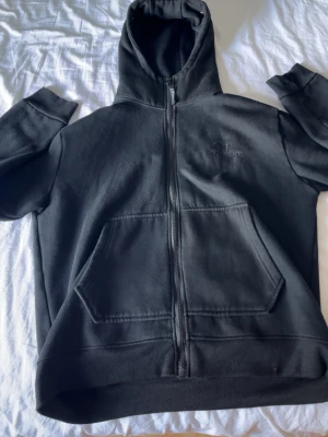 Svart hoodie med glasögon från C.P. Company - Säljer en svart hoodie från C.P. Company med dragkedja, stor ficka framtill och ikoniska inbyggda glasögon i huvan. Broderad logga på bröstet och coola detaljer i metall runt huvan. Perfekt för dig som gillar streetwear och unika plagg.