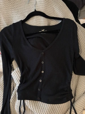 Svart ribbad topp från Hollister - Snygg svart ribbad topp från Hollister Must-Have Collection. Modellen är en baby tee med långa ärmar, knappar framtill och dragsko i sidorna för en trendig look. Perfekt att styla med jeans eller kjol för en cool vardagsoutfit.